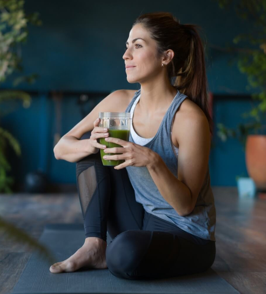 Fit woman in workout attire sitting on a yoga mat, holding a green smoothie and relaxing in a peaceful wellness studio